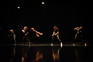 A group of dancers performing on a dimly lit stage, their movements captured in mid-action with dramatic lighting highlighting their forms against the dark background.