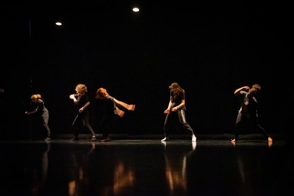 A group of dancers performing on a dimly lit stage, their movements captured in mid-action with dramatic lighting highlighting their forms against the dark background.