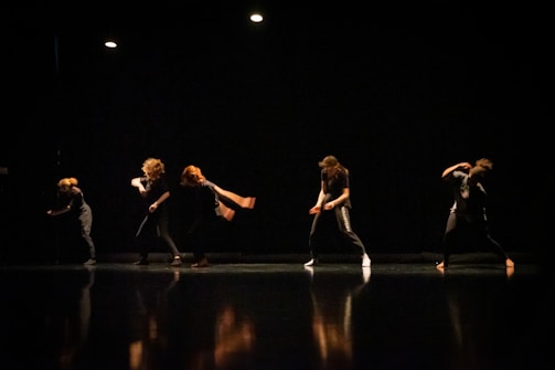 A group of dancers performing on a dimly lit stage, their movements captured in mid-action with dramatic lighting highlighting their forms against the dark background.