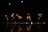 A group of dancers performing on a dimly lit stage, their movements captured in mid-action with dramatic lighting highlighting their forms against the dark background.