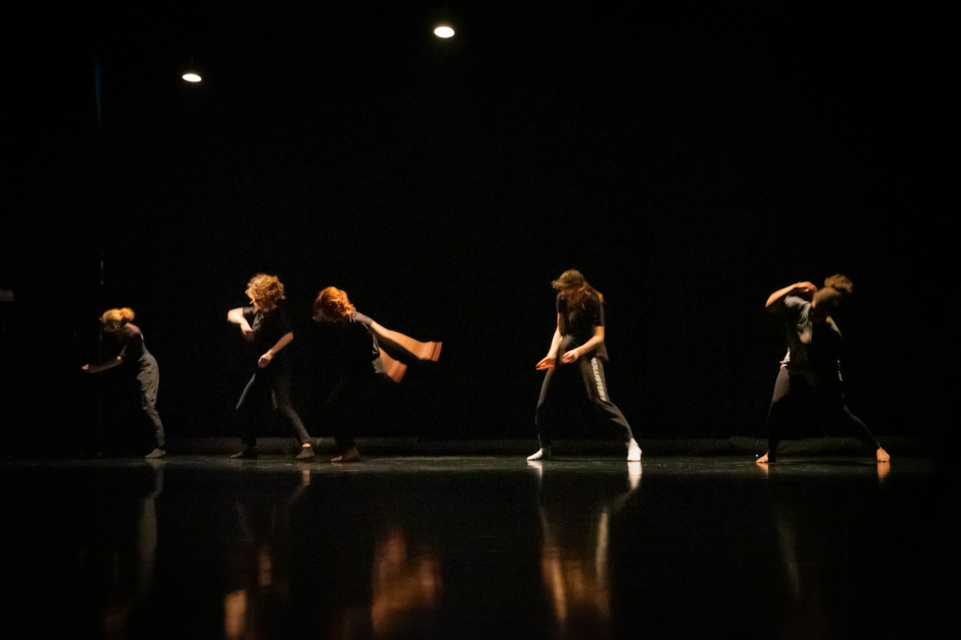 A group of dancers performing on a dimly lit stage, their movements captured in mid-action with dramatic lighting highlighting their forms against the dark background.