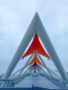 A modern architectural structure featuring a covered pedestrian bridge with geometric elements. Triangular supports and colorful fabric canopies in red, orange, and purple shades create a striking visual pattern. The bridge spans a wide walkway with glass railings and extends into the distance. The sky is overcast, adding an element of calm to the scene.