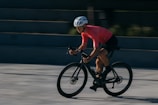 A cyclist wearing a bright pink jersey and black shorts is riding a bike rapidly, leaning forward with focus and intensity. The cyclist is wearing a white helmet and sunglasses. The background is blurred, suggesting a sense of speed and motion.