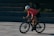 A cyclist wearing a bright pink jersey and black shorts is riding a bike rapidly, leaning forward with focus and intensity. The cyclist is wearing a white helmet and sunglasses. The background is blurred, suggesting a sense of speed and motion.