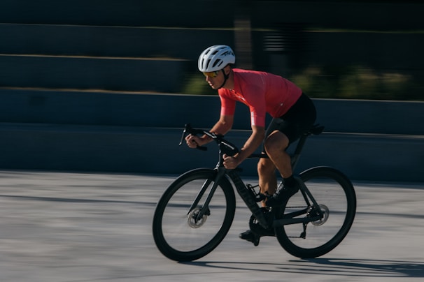 A cyclist wearing a bright pink jersey and black shorts is riding a bike rapidly, leaning forward with focus and intensity. The cyclist is wearing a white helmet and sunglasses. The background is blurred, suggesting a sense of speed and motion.