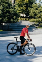 A person wearing a red cycling jersey and black shorts is standing next to a black bicycle. The person is also wearing a white helmet and sunglasses. The background includes some greenery with trees and concrete steps.