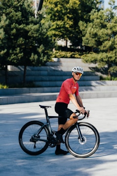 A person wearing a red cycling jersey and black shorts is standing next to a black bicycle. The person is also wearing a white helmet and sunglasses. The background includes some greenery with trees and concrete steps.