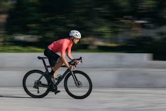 A cyclist wearing a white helmet and a pink shirt rides a sleek black bicycle at high speed. The background is blurred, indicating motion, with a mix of greenery and a light-colored wall.