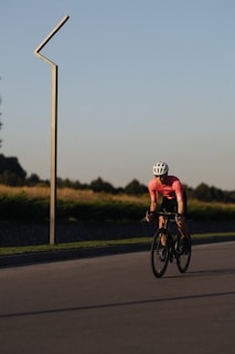 A cyclist wearing a pink sports jersey and white helmet is riding a bicycle along a paved road. The setting appears to be outdoors during a clear day, evidenced by the open landscape and clear sky. There is a vertical metal structure on the left side of the road. The background includes green bushes and some trees in the distance.