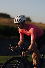 A cyclist is riding a bicycle wearing a white helmet and a pink cycling jersey. The cyclist is focused, and the background features a blurred landscape of greenery and a hill, likely taken during a sunset or sunrise.
