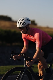 A cyclist is riding a bicycle wearing a white helmet and a pink cycling jersey. The cyclist is focused, and the background features a blurred landscape of greenery and a hill, likely taken during a sunset or sunrise.