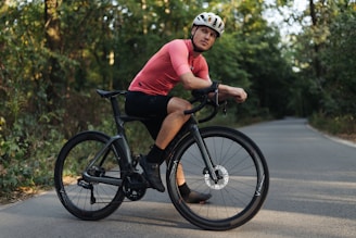 A cyclist is resting on a black road bike in a peaceful wooded area. He is wearing a bright pink cycling shirt, black shorts, black shoes, and a white helmet. The road curves gently and is surrounded by dense green foliage, suggesting a serene and natural setting.
