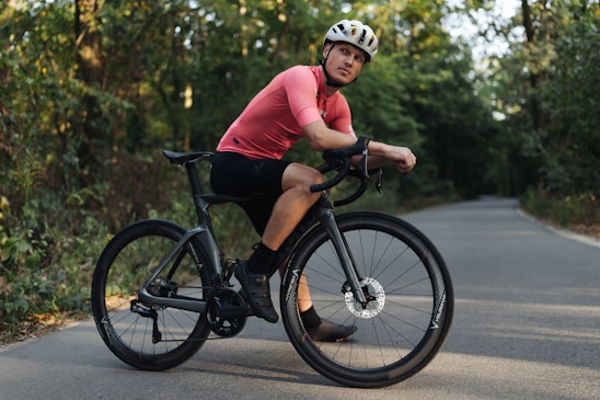 A cyclist is resting on a black road bike in a peaceful wooded area. He is wearing a bright pink cycling shirt, black shorts, black shoes, and a white helmet. The road curves gently and is surrounded by dense green foliage, suggesting a serene and natural setting.