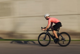 A cyclist wearing a bright pink jersey and a white helmet rides a bicycle at high speed. The background is blurred, emphasizing motion and the dynamic nature of the scene.