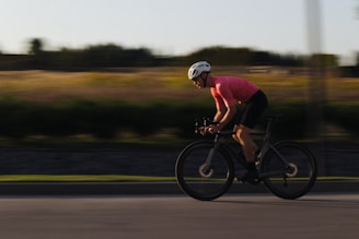 A cyclist wearing a pink jersey and a white helmet rides quickly along a road. The background appears blurred, suggesting motion, with fields and trees in the distance.