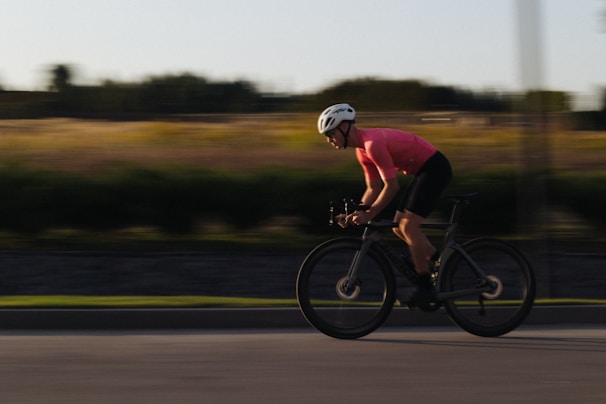 A cyclist wearing a pink jersey and a white helmet rides quickly along a road. The background appears blurred, suggesting motion, with fields and trees in the distance.