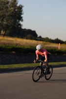 A cyclist wearing a pink jersey and white helmet rides a bicycle on a smooth road in a rural setting. The background features grassy fields and a few trees under a clear blue sky, suggesting a peaceful, natural environment.