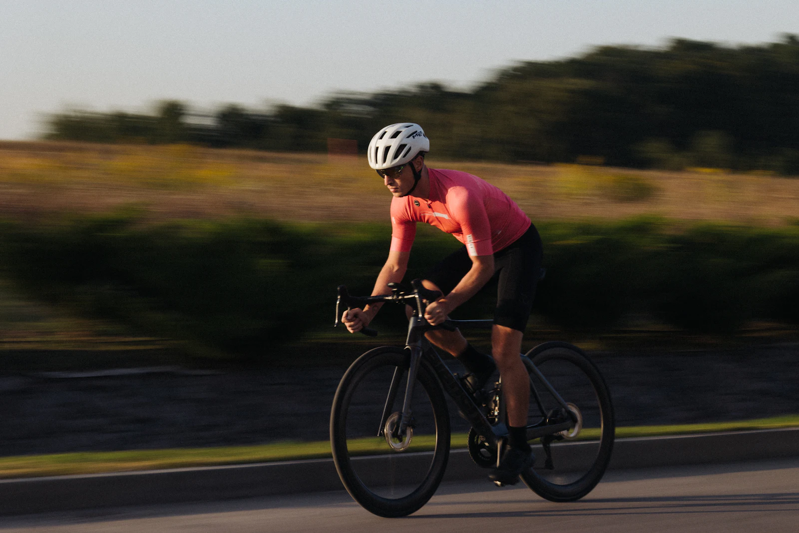 A road cyclist in a pink jersey and white helmet riding fast along a road in low evening sun, with grass and trees blurred behind