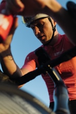 A cyclist wearing a helmet and a pink cycling jersey is captured from a low angle perspective. The focus is on his intense expression as he appears to be in action, possibly during a race or a strenuous ride. His hand is reaching towards a red water bottle, and there's a bike handlebar prominently visible.