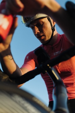 A cyclist wearing a helmet and a pink cycling jersey is captured from a low angle perspective. The focus is on his intense expression as he appears to be in action, possibly during a race or a strenuous ride. His hand is reaching towards a red water bottle, and there's a bike handlebar prominently visible.