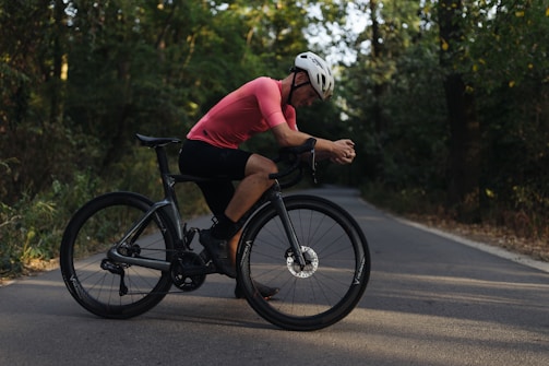 A cyclist in a pink jersey and white helmet is resting on his black bicycle. He is on a paved road surrounded by lush greenery, with trees and bushes lining the route. The lighting suggests a serene late afternoon setting.