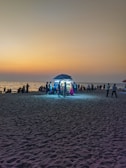 A beach scene at sunset with a street food stall under an umbrella illuminated by bright lights. Several people are gathered around the stall, and more can be seen along the shoreline. The sky is a gradient of warm colors and the sand captures the ambient light.