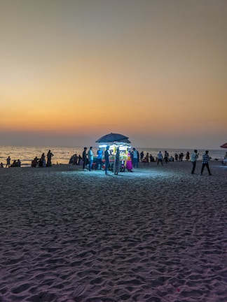 A beach scene at sunset with a street food stall under an umbrella illuminated by bright lights. Several people are gathered around the stall, and more can be seen along the shoreline. The sky is a gradient of warm colors and the sand captures the ambient light.