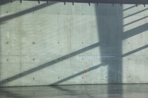 Shadows stretch across a plain concrete wall, creating diagonal lines. The surface is smooth and dotted with small circular holes, providing a minimalist and industrial aesthetic. The bottom reflection on the polished floor adds depth to the scene.