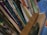 A collection of books and folders stacked vertically in a shelf or storage area. The book titles are visible, indicating subjects such as mathematics, genetics, biotechnology, and English, aimed at senior secondary and undergraduate levels. A blue container and a brown envelope are also present alongside the books.
