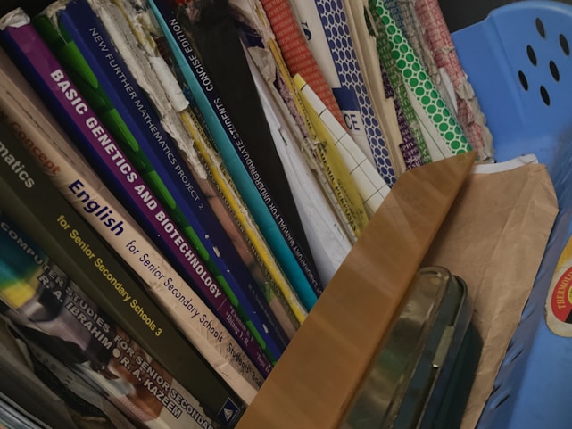 A collection of books and folders stacked vertically in a shelf or storage area. The book titles are visible, indicating subjects such as mathematics, genetics, biotechnology, and English, aimed at senior secondary and undergraduate levels. A blue container and a brown envelope are also present alongside the books.
