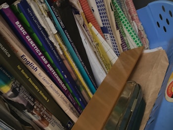 A collection of books and folders stacked vertically in a shelf or storage area. The book titles are visible, indicating subjects such as mathematics, genetics, biotechnology, and English, aimed at senior secondary and undergraduate levels. A blue container and a brown envelope are also present alongside the books.
