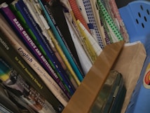 A collection of books and folders stacked vertically in a shelf or storage area. The book titles are visible, indicating subjects such as mathematics, genetics, biotechnology, and English, aimed at senior secondary and undergraduate levels. A blue container and a brown envelope are also present alongside the books.