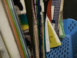 A collection of textbooks and notebooks in various colors with visible spines. Some of the books include subjects like English and sciences. The materials are organized upright in a blue plastic basket.