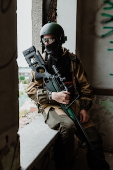 A person dressed in tactical gear is sitting by a window in a rundown building. The individual is wearing a helmet and goggles, with a face covering that obscures their expression. Their attire includes a brown jacket and camouflage pants, and they are holding a large firearm. The setting suggests an urban environment, possibly during a conflict or training exercise.