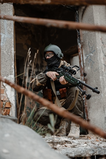 A person in full tactical gear, including a helmet and face covering, is crouched in a derelict urban setting. They are holding a sniper rifle and appear to be on alert. The background shows a damaged building with visible rusted metal bars and crumbling concrete.