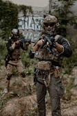 Two individuals are dressed in military-style gear, holding rifles while standing in a grassy, outdoor area. They are wearing helmets and tactical vests, and one person is aiming forward. Graffiti can be seen on a wall behind them.