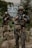Two individuals are dressed in military-style gear, holding rifles while standing in a grassy, outdoor area. They are wearing helmets and tactical vests, and one person is aiming forward. Graffiti can be seen on a wall behind them.