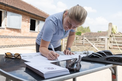 A person with blonde hair is leaning over a table, signing or writing on a document with a pen. The table is outdoors, with a brick building and a wooden fence in the background. Various objects are on the table, including sunglasses, a coiled rope, and stacks of paper.