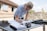 A person with blonde hair is leaning over a table, signing or writing on a document with a pen. The table is outdoors, with a brick building and a wooden fence in the background. Various objects are on the table, including sunglasses, a coiled rope, and stacks of paper.
