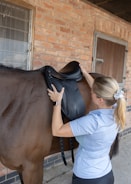 A person is adjusting a black saddle on the back of a brown horse. The setting is an outdoor stable with a brick wall and a wooden door in the background. The individual is wearing a light blue shirt and has a smartwatch on their wrist.