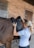 A person is adjusting a black saddle on the back of a brown horse. The setting is an outdoor stable with a brick wall and a wooden door in the background. The individual is wearing a light blue shirt and has a smartwatch on their wrist.
