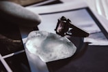 A close-up of an ornate ring with a dark gemstone placed on a translucent, irregularly shaped crystal. Nearby lies a smooth, oval stone with a matte texture. The background is composed of blurred photographs or prints, adding an artistic and enigmatic atmosphere to the composition.