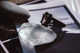 A close-up of an ornate ring with a dark gemstone placed on a translucent, irregularly shaped crystal. Nearby lies a smooth, oval stone with a matte texture. The background is composed of blurred photographs or prints, adding an artistic and enigmatic atmosphere to the composition.