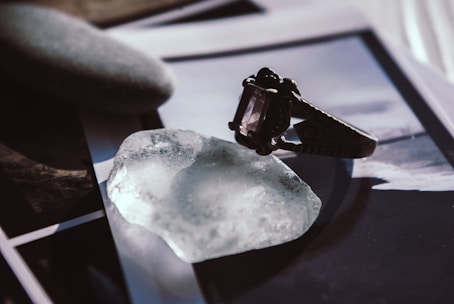 A close-up of an ornate ring with a dark gemstone placed on a translucent, irregularly shaped crystal. Nearby lies a smooth, oval stone with a matte texture. The background is composed of blurred photographs or prints, adding an artistic and enigmatic atmosphere to the composition.