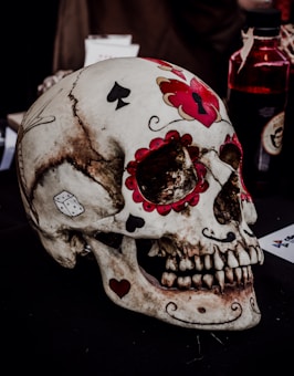 A skull is decorated with colorful patterns and symbols, including playing card suits and dice. The skull has intricate red, black, and white designs that resemble Day of the Dead motifs. It is placed on a dark surface with a blurred background, including a bottle and some papers.