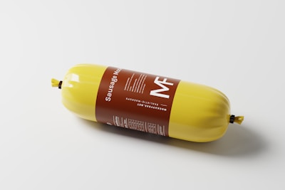 A cylindrical sausage wrapped in bright yellow packaging, with a brown label that includes text and nutritional information. The packaging is sealed at both ends with yellow twists.