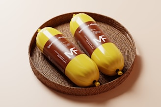 Two yellow packaged sausage mock-ups placed in a round woven basket on a light surface. The packaging features brown labels with text and the letters 'MF'.