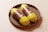 Two yellow packaged sausage mock-ups placed in a round woven basket on a light surface. The packaging features brown labels with text and the letters 'MF'.