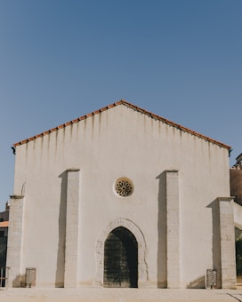 A minimalist church facade with clean lines and neutral colors. The structure features a triangular roof with red tiles and vertical pillars. A round window with a decorative design is situated above a large wooden arched door.