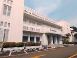 A three-story white building with an art deco architectural style, featuring horizontal lines and large windows. The facade displays the name 'Bappenas' and 'Badan Perencanaan Pembangunan Nasional'. Well-maintained shrubs and potted plants line the building exterior. An empty parking lot with marked spaces is in the foreground, bordered by yellow lines. A clear blue sky is visible above.